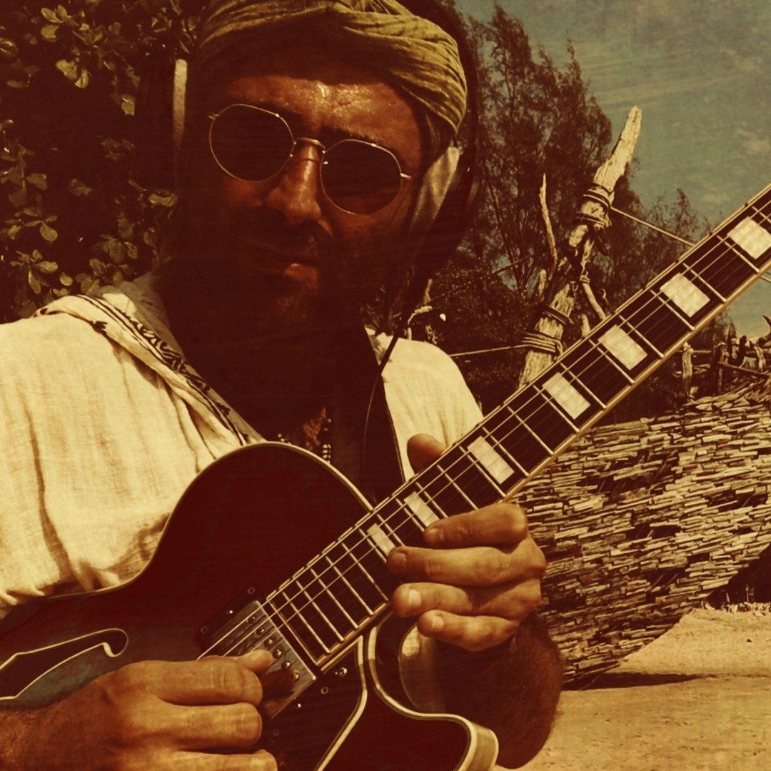 Constantine Cheimarios playing his archtop guitar, in round sunglasses and turban
